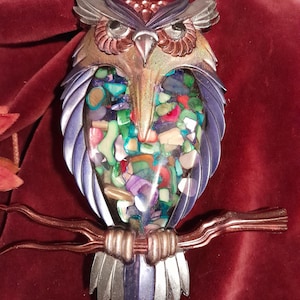 May include: A decorative owl figurine made of metal and colorful mosaic pieces. The owl has a silver and bronze head, with a body filled with various colored mosaic pieces. It is perched on a bronze-colored branch, set against a red background.