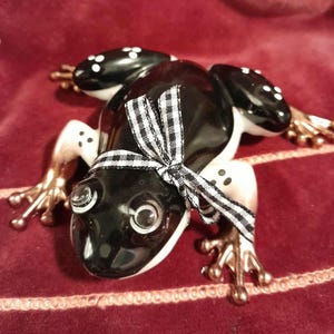 May include: A ceramic frog figurine with a glossy black body, white spots, and bronze-colored feet. It has a black and white checkered bow around its neck. The frog is lying on a red surface.