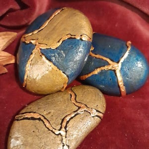 May include: Three painted stones with a Kintsugi-style design. The stones are blue, gold, and tan, with gold and black lines. The stones are on a red surface.