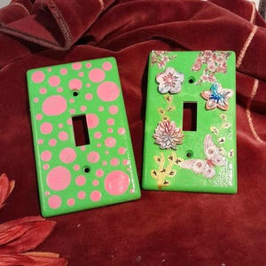 May include: Two decorative light switch covers. One is lime green with pink polka dots. The other is lime green with floral and butterfly designs. Both have a rectangular opening for the switch.