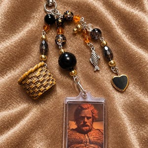 May include: A keyring featuring a heart-shaped clasp, a rectangular frame containing a portrait of a bearded man, a miniature woven basket charm, a silver fish charm, and a black heart charm, accented with black, gold, and amber beads.