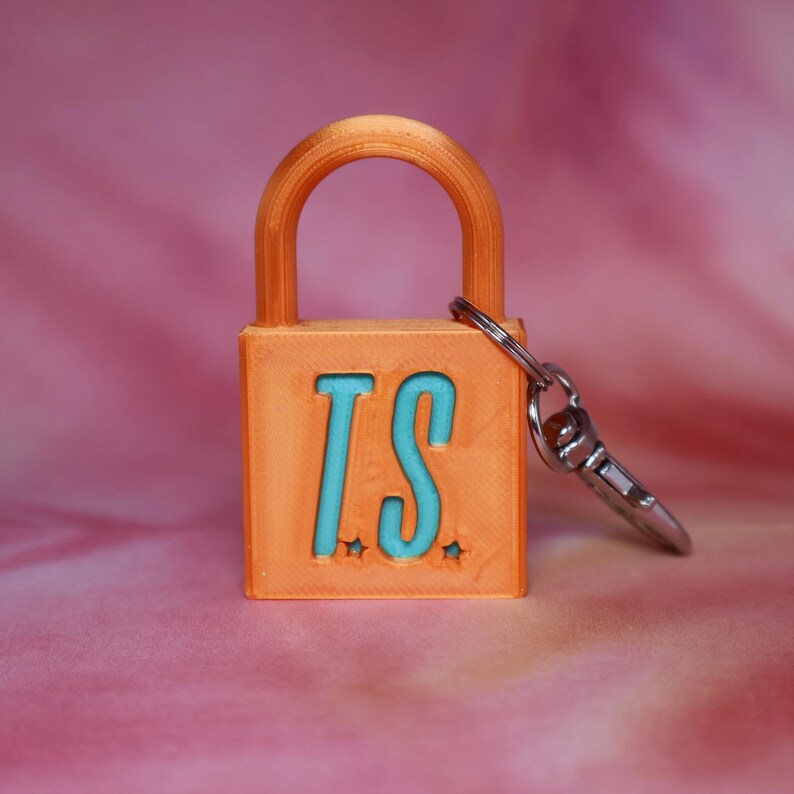 May include: An orange padlock keychain featuring the letters T.S. in a bright turquoise blue color on the front face. The padlock has a rounded shackle and a silver-toned metal lobster clasp attached to the side, with small star accents near the letters.