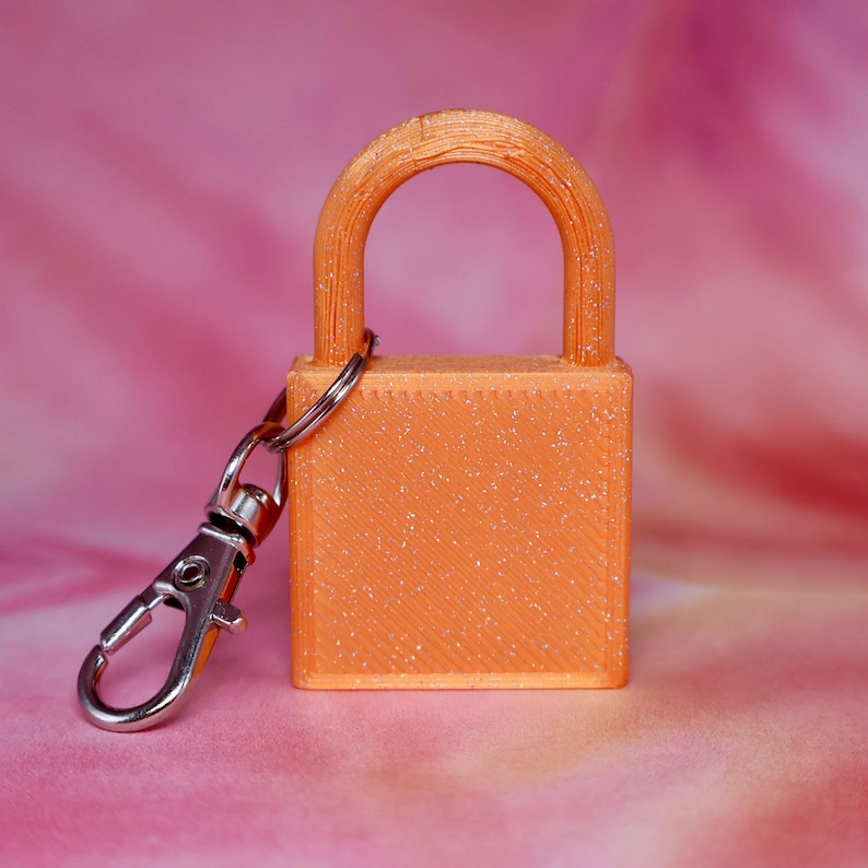 May include: A bright orange padlock keychain with a glittery finish and a silver-toned metal lobster clasp attached to the side. The padlock features a rectangular body and a rounded shackle, showing a textured surface from the manufacturing process.