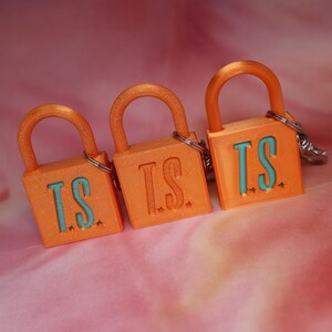 May include: Three orange padlock keychains with a glittery finish. Each padlock features the letters T.S. in a light blue color on the front, accompanied by small star shapes. A silver metal ring is attached to the top of each orange padlock.