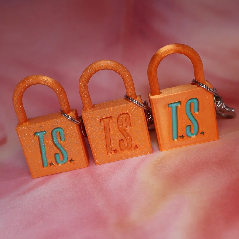 May include: Three orange padlock keychains with a glittery finish. Each padlock features the letters T.S. in a light blue color on the front, accompanied by small star shapes. A silver metal ring is attached to the top of each orange padlock.