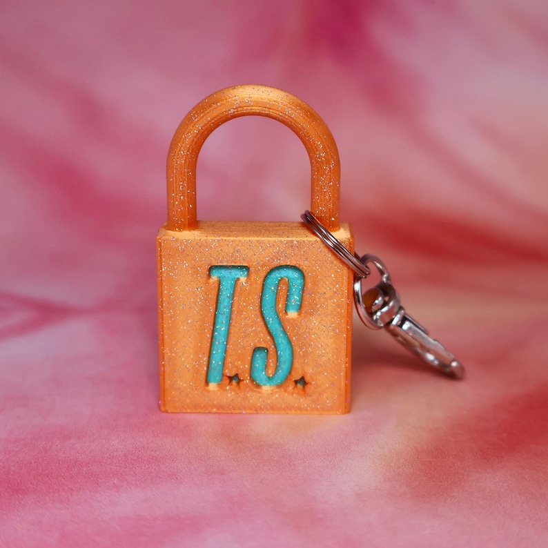 May include: An orange glitter padlock keychain featuring the letters TS in a bright teal color on the front face. The padlock has a rounded shackle and includes a silver-toned metal lobster clasp attached to the side for easy hanging.