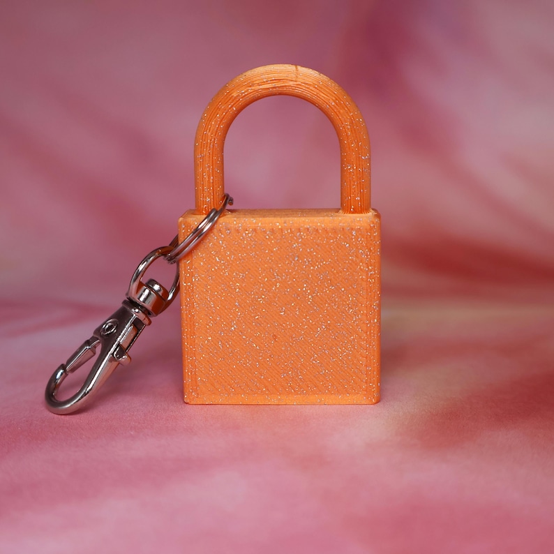 May include: A bright orange padlock keychain with a visible glittery texture throughout the surface. The padlock features a standard U-shaped shackle and a rectangular body, attached to a silver-toned metal lobster clasp via a small metal ring.