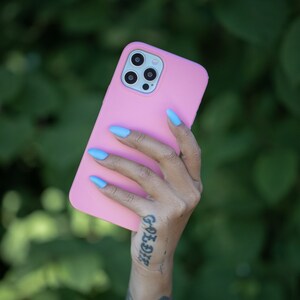 May include: A pink phone case with a camera lens cutout. The case is being held by a hand with blue nail polish and a tattoo on the wrist.