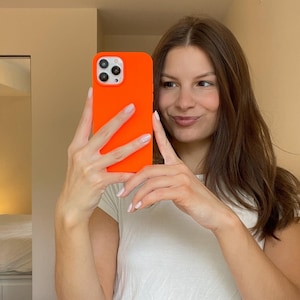 May include: A woman is holding a bright orange iPhone case in front of a mirror. The case has a raised camera bump and a matte finish.