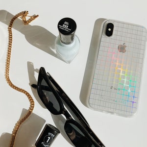 Könnte beinhalten: Eine flache Anordnung verschiedener Gegenstände, darunter eine transparente Handyhülle mit einem Regenbogen-Gittermuster, eine schwarze Cat-Eye-Sonnenbrille, eine Flasche hellblauen Nagellacks und eine Flasche klaren Nagellacks mit schwarzem Deckel und dem Text "STEP 1 ETAPE" auf dem Etikett.