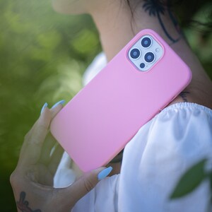 May include: A pink silicone phone case with a camera lens cutout. The case is being held by a person with blue nail polish.