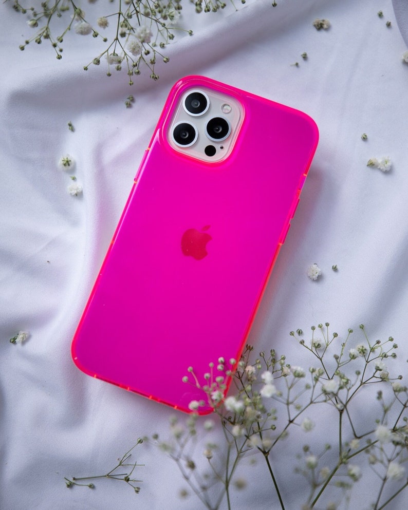 May include: A bright pink phone case with a clear back and a black Apple logo. The case has a raised camera bump with three black camera lenses.