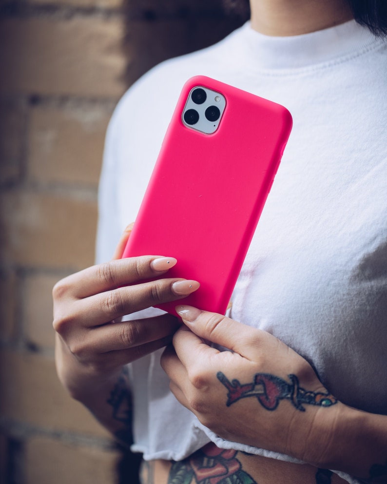 May include: A bright pink smartphone case with a triple-lens camera is held against a white shirt. The case is a solid, vibrant color, and the phone's camera lenses are visible. The background is a brick wall.