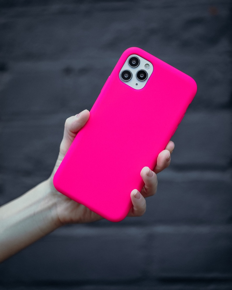 May include: A bright pink phone case, held in a hand, is the focus of this image. The case is a solid, vibrant pink, and appears to be made of a smooth material. The phone's camera lenses are visible. The background is a dark, textured surface.