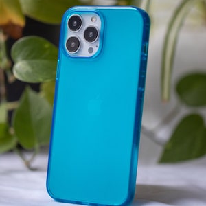 May include: A turquoise phone case with a clear, transparent design. The case has a smooth surface and a protective bumper around the edges. The phone case is shown against a backdrop of green foliage.