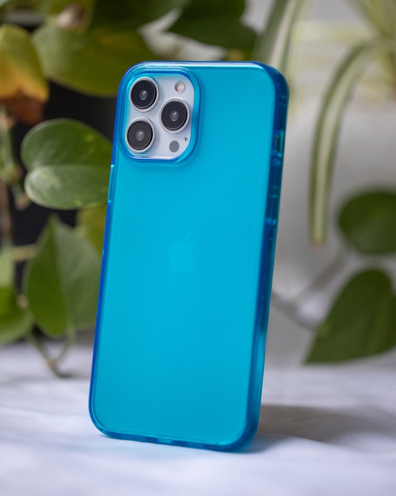May include: A turquoise phone case with a clear, transparent design. The case has a smooth surface and a protective bumper around the edges. The phone case is shown against a backdrop of green foliage.