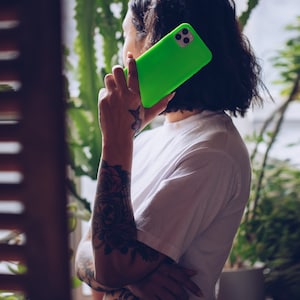 K&ouml;nnte beinhalten: Eine Person mit T&auml;towierungen am Arm h&auml;lt eine leuchtend gr&uuml;ne Handyh&uuml;lle mit einem schwarzen Kameraobjektiv. Die Person tr&auml;gt ein wei&szlig;es T-Shirt und eine blaue Jeans.