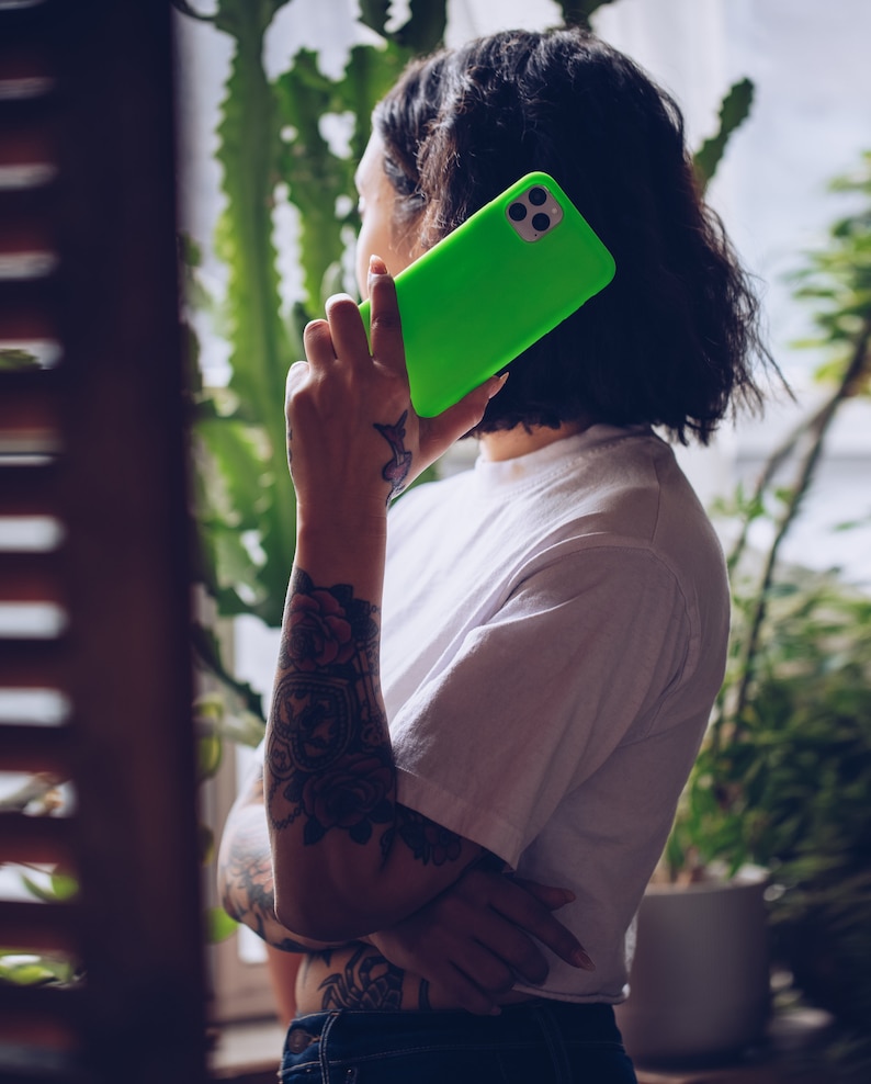 K&ouml;nnte beinhalten: Eine Person mit T&auml;towierungen am Arm h&auml;lt eine leuchtend gr&uuml;ne Handyh&uuml;lle mit einem schwarzen Kameraobjektiv. Die Person tr&auml;gt ein wei&szlig;es T-Shirt und eine blaue Jeans.