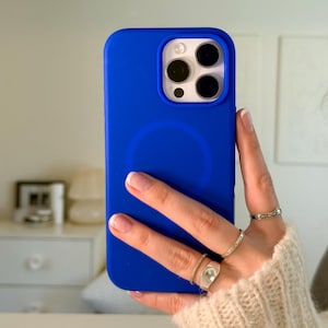 Op de afbeelding: Een felblauwe telefoonhoes met een cirkelvormig ontwerp op de achterkant. De hoes wordt vastgehouden in een hand met meerdere ringen. De cameralenzen van de telefoon zijn zichtbaar.