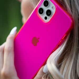 May include: A bright pink phone case with a clear back and a black Apple logo. The case has a raised camera bump with three black camera lenses.