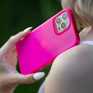 May include: A bright pink phone case with a clear back and a raised camera bump. The case is made of a flexible material and has a smooth finish.