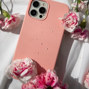 May include: A pink silicone phone case with a clear camera lens cover. Water droplets are visible on the case. The case is surrounded by pink carnations.