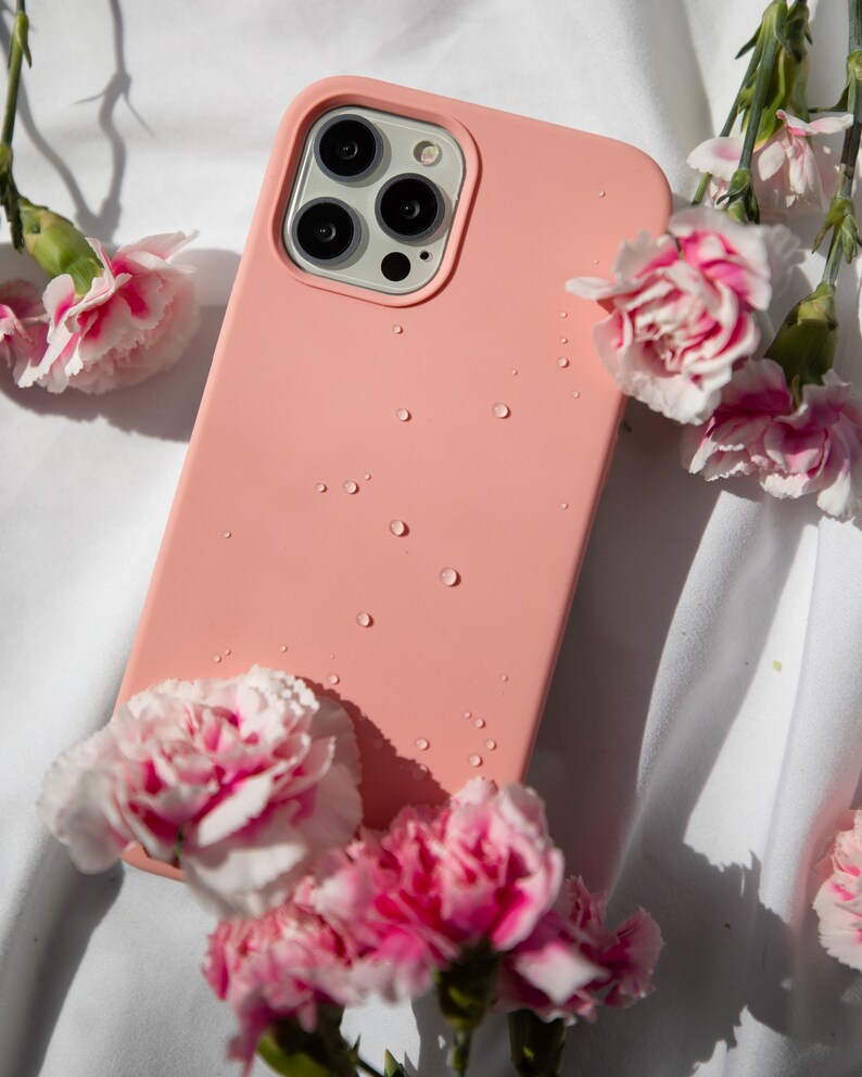 May include: A pink silicone phone case with a clear camera lens cover. Water droplets are visible on the case. The case is surrounded by pink carnations.