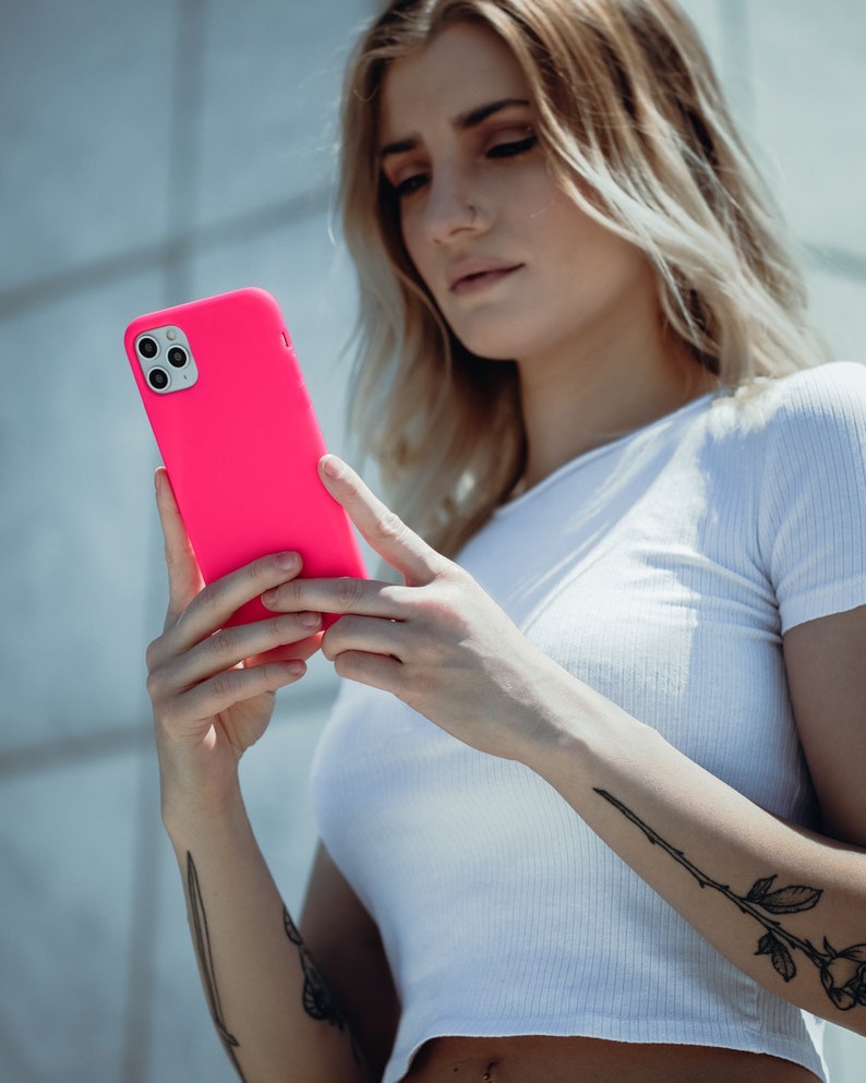 May include: A bright pink smartphone case. The case is a vibrant, solid color and appears to be made of a smooth material. The phone case is held in front of a person wearing a white shirt.