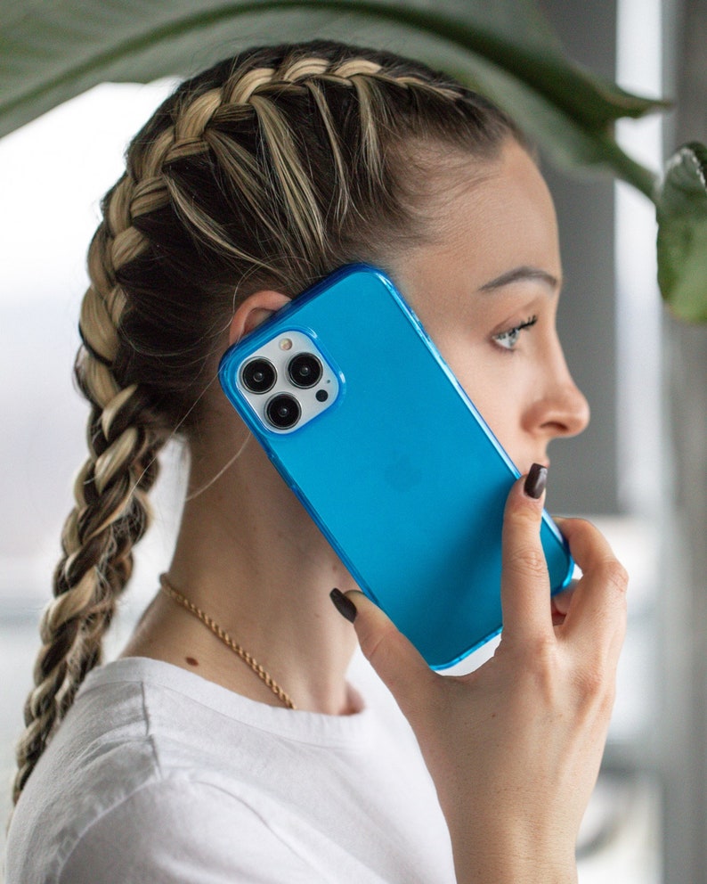 May include: A bright blue smartphone case with a clear border. The phone case is held up to the ear. The phone has three camera lenses. The person is wearing a white shirt and has a braided hairstyle.