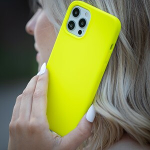May include: A bright yellow phone case with a black camera lens and a white border. The case is held in a person's hand.