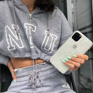 以下が含まれることがあります： 真珠のような虹色のデザインの透明な携帯電話ケース。ケースは、小さく、白く、虹色のキラキラしたようなもので飾られています。ケースは、薄い青色のマニキュアをした人によって持たれています。