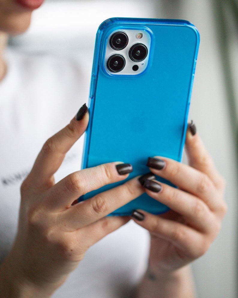 May include: A bright blue phone case with a clear border, held in hands. The case covers a phone with three camera lenses. The phone case is a vibrant, solid color, and the clear border adds a sleek design element.
