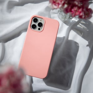 May include: A pink silicone phone case with a black camera lens cutout. The case is lying on a white fabric surface.