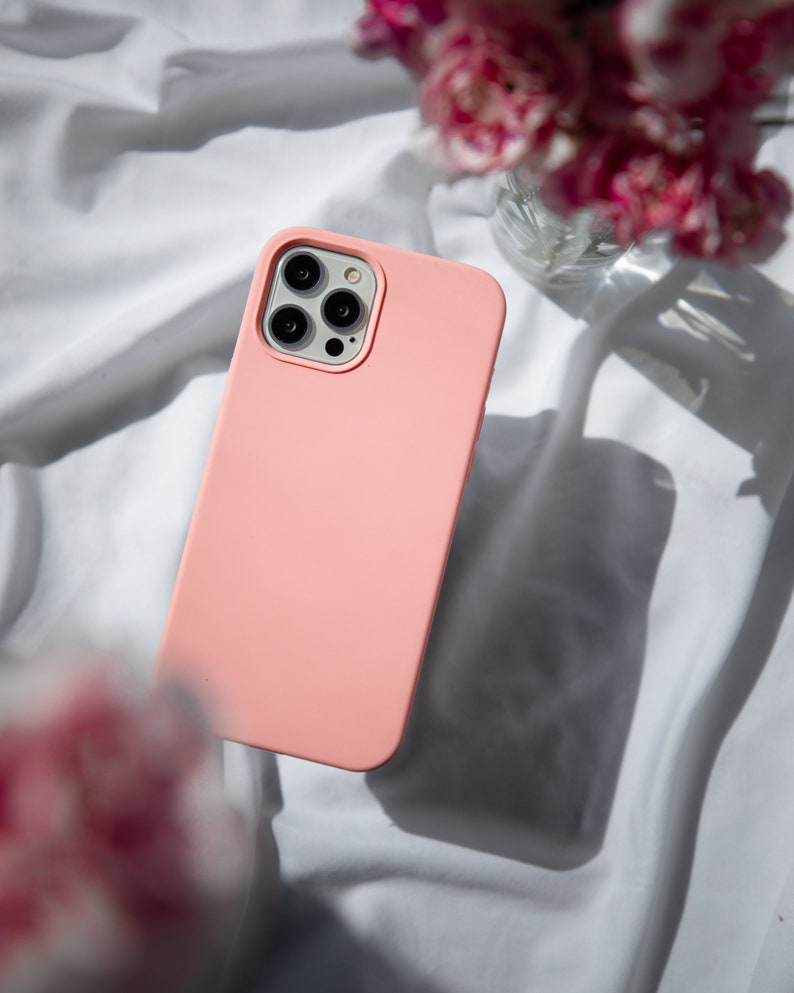 May include: A pink silicone phone case with a black camera lens cutout. The case is lying on a white fabric surface.