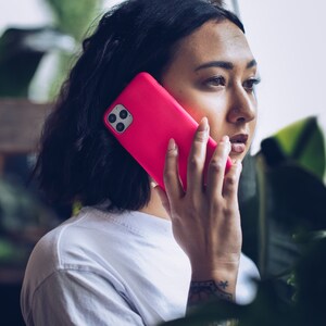 May include: A bright pink smartphone case with a triple-lens camera is held up to the ear. The person is wearing a white t-shirt. The background is blurred with green foliage.