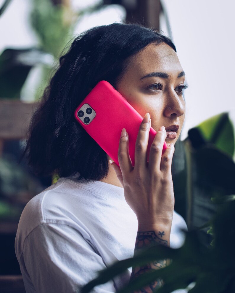 May include: A bright pink smartphone case with a triple-lens camera is held up to the ear. The person is wearing a white t-shirt. The background is blurred with green foliage.