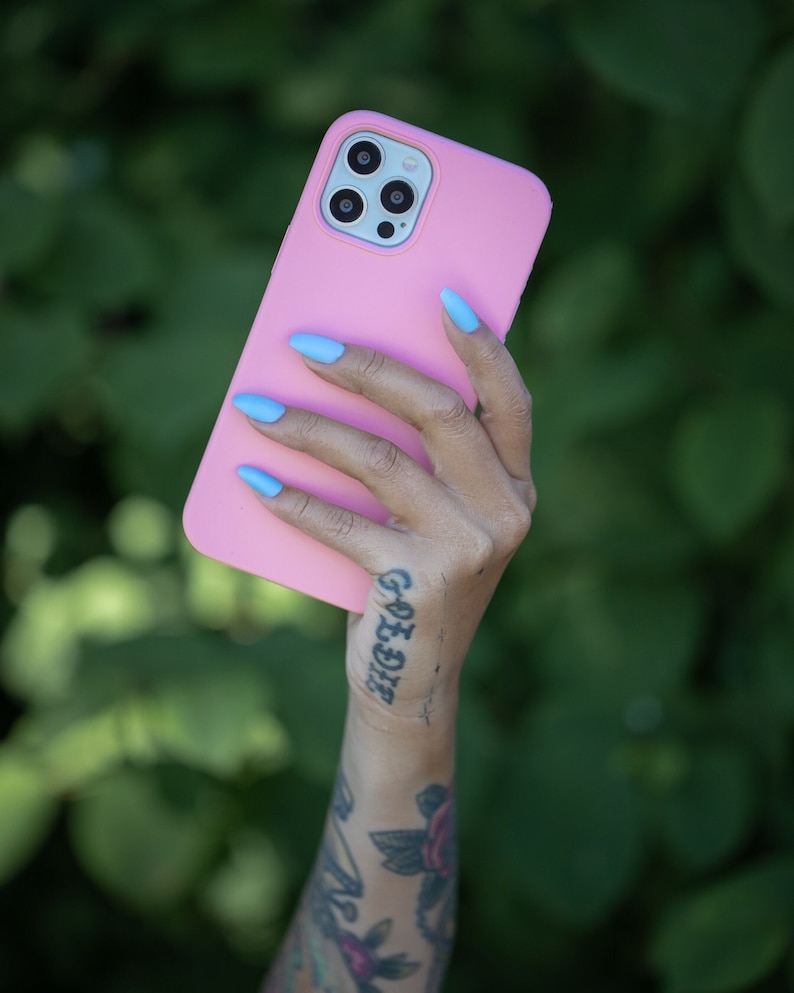 May include: A pink phone case with a camera lens cutout. The case is being held by a hand with blue nail polish and a tattoo on the wrist.