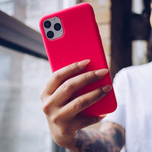 May include: A bright pink smartphone case with a triple-lens camera is held up. The case is a solid, vibrant color. The phone is held in front of a window, with a person's arm and hand visible.