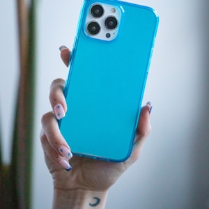 May include: A bright turquoise phone case with a clear border. The phone case is held in a hand with manicured nails. The phone has three camera lenses.