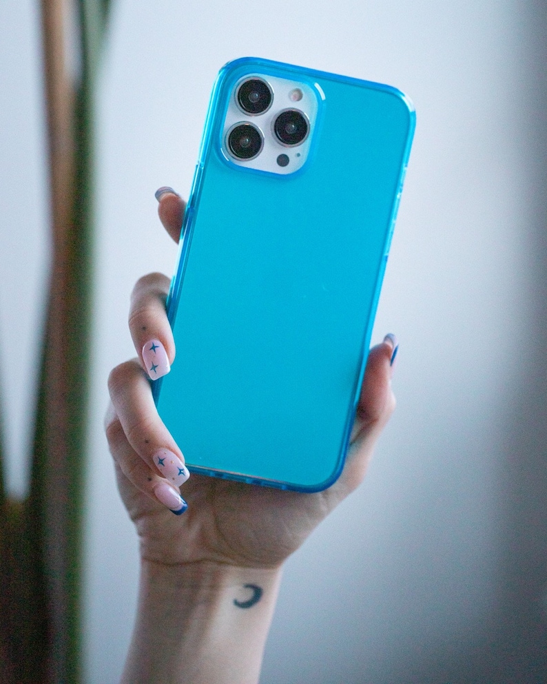 May include: A bright turquoise phone case with a clear border. The phone case is held in a hand with manicured nails. The phone has three camera lenses.