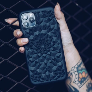 May include: A dark grey phone case featuring a geometric design and a central floral motif. The case has a textured surface and is held in a hand.