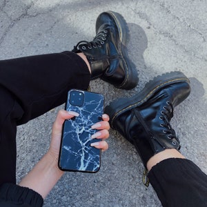 May include: A black and white marble phone case with a white background and black veins. The case is being held by a person wearing black pants and black boots.
