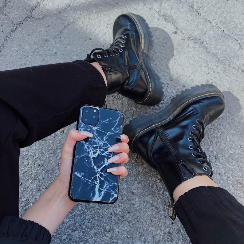 May include: A black and white marble phone case with a white background and black veins. The case is being held by a person wearing black pants and black boots.