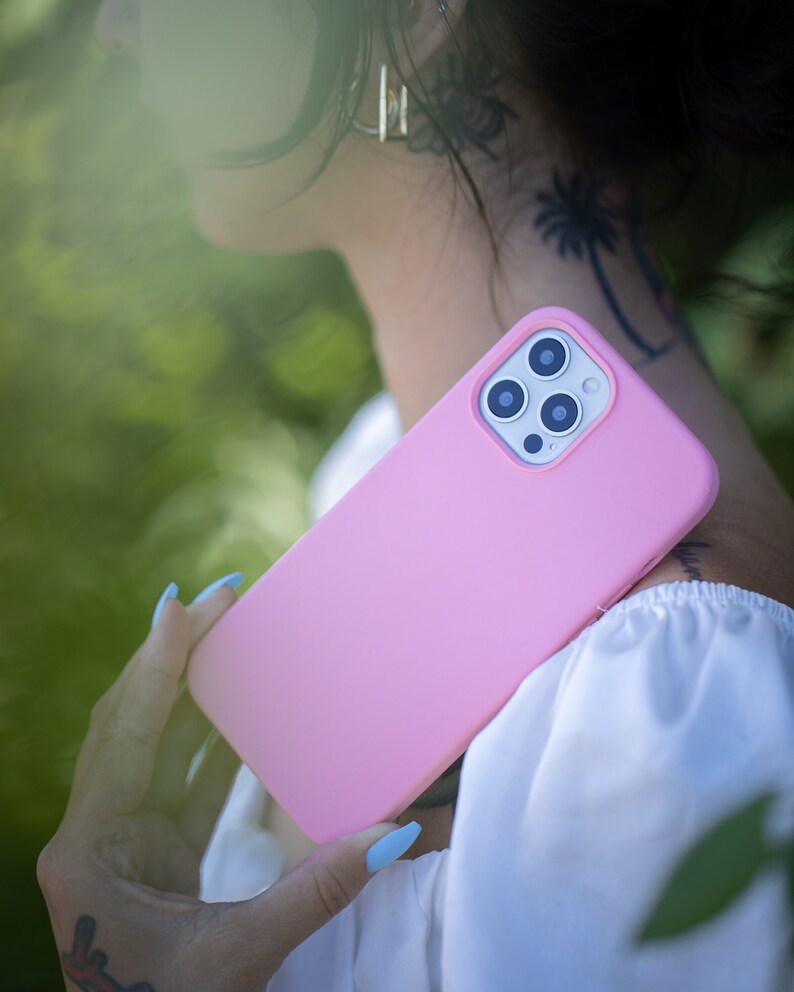 May include: A pink silicone phone case with a camera lens cutout. The case is being held by a person with blue nail polish.