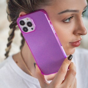 May include: A purple phone case with a clear back and a raised camera bump. The case is made of a flexible material and has a smooth finish.