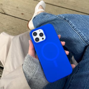 Op de afbeelding: Een felblauwe telefoonhoes met een cirkelvormig ontwerp aan de achterkant. De hoes wordt in de hand van een persoon gehouden, met de camera van de telefoon zichtbaar. De achtergrond omvat een spijkerbroek en een houten oppervlak.