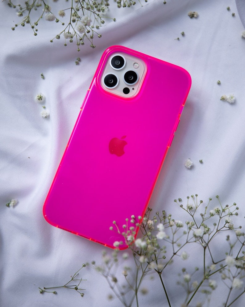 May include: A bright pink phone case with a clear camera lens cover and an Apple logo. The case is lying on a white surface with small white flowers.