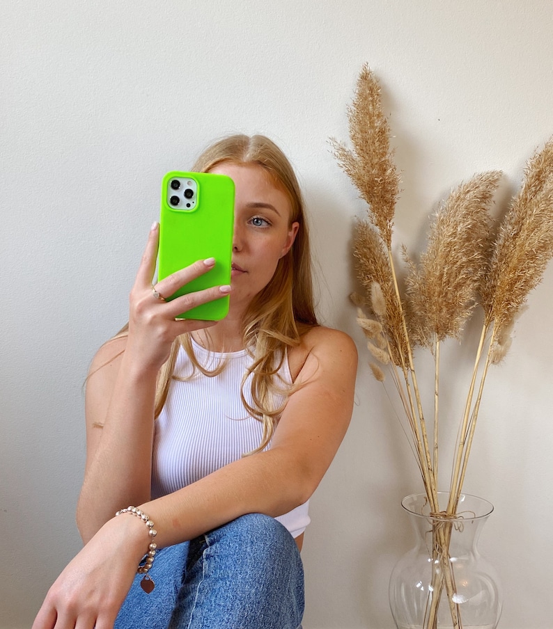 K&ouml;nnte beinhalten: Eine Frau h&auml;lt eine leuchtend gr&uuml;ne Handyh&uuml;lle vor ihrem Gesicht. Die H&uuml;lle hat einen schwarzen Ausschnitt f&uuml;r die Kamera. Sie tr&auml;gt ein wei&szlig;es Tanktop und eine blaue Jeans.