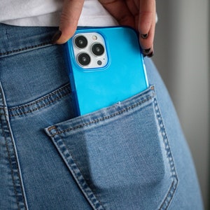 May include: A bright blue smartphone in a denim jeans pocket. The phone has a clear case and a triple-lens camera. The jeans are a light blue wash, and the pocket is visible.
