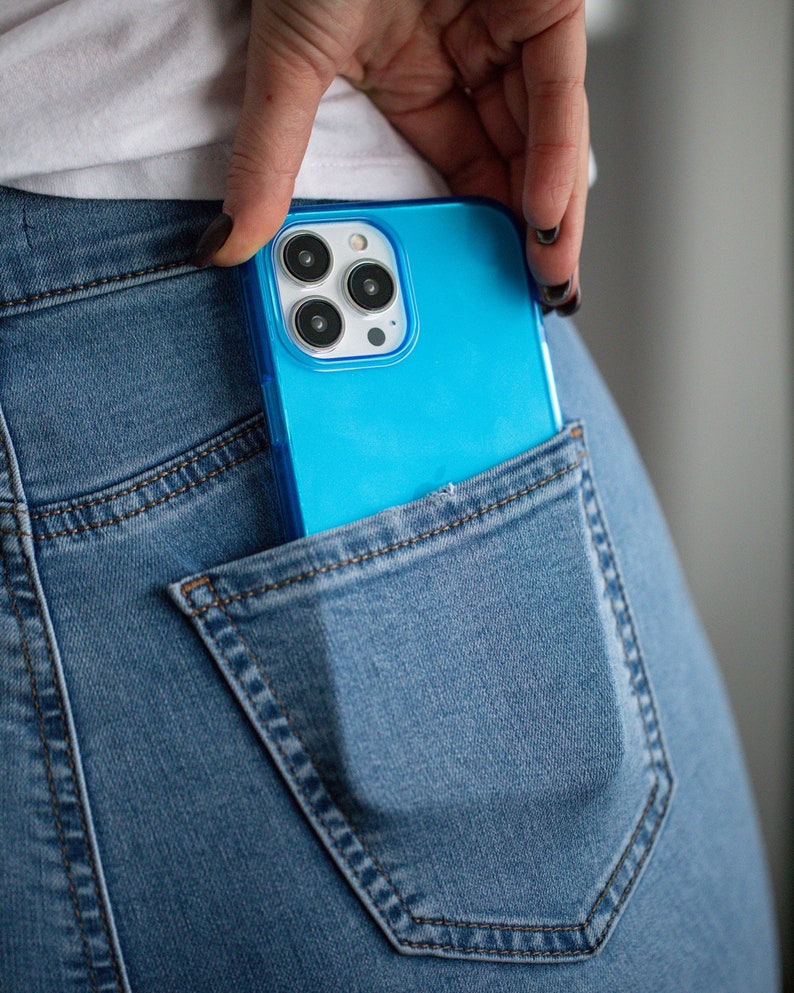 May include: A bright blue smartphone in a denim jeans pocket. The phone has a clear case and a triple-lens camera. The jeans are a light blue wash, and the pocket is visible.